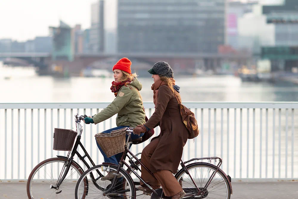 Two women cycling side by side over Knippelsbro with the harbour behind them