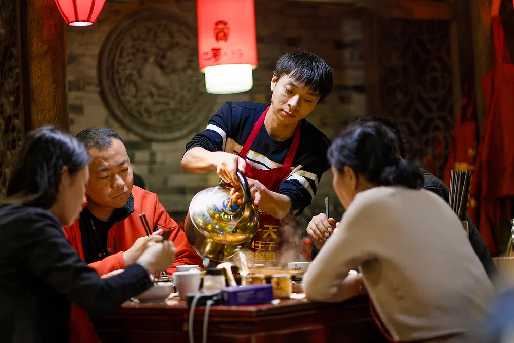 Family eating hot pot while a waiter pours from a brass teapot in Chengdu