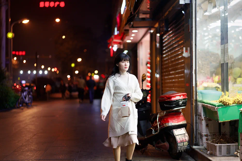 Woman in a white coat walking down a lit shopping street at night