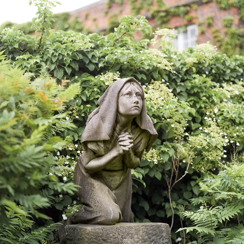 Weathered statue of a kneeling hooded woman praying among green leaves