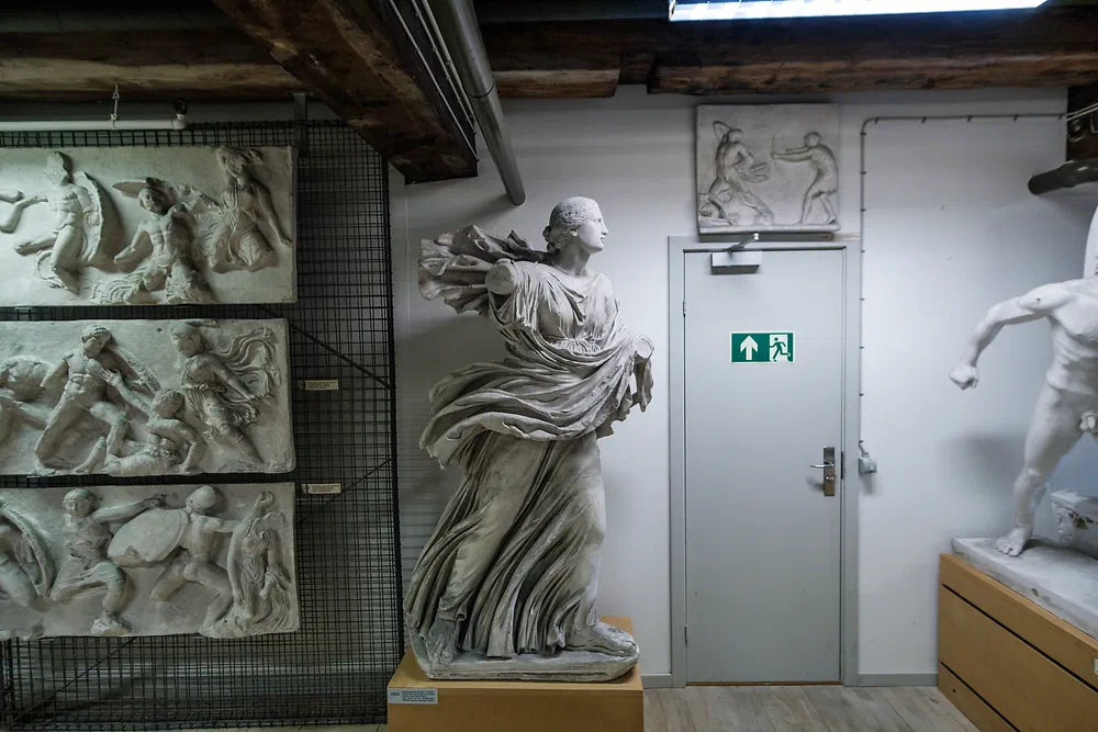 Plaster cast of a female statue in a white storage room with reliefs on the walls