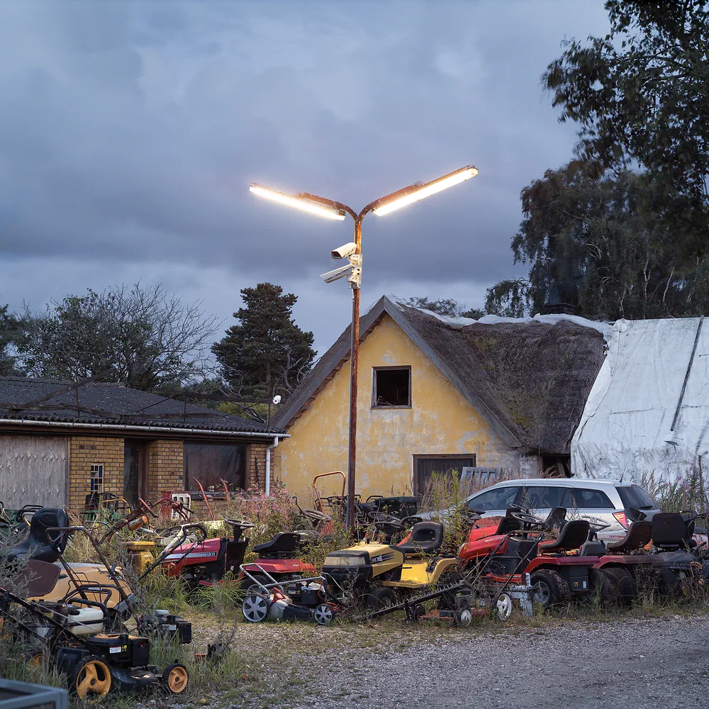 Rows of used lawnmowers in front of a thatched yellow house under a street lamp in Vig