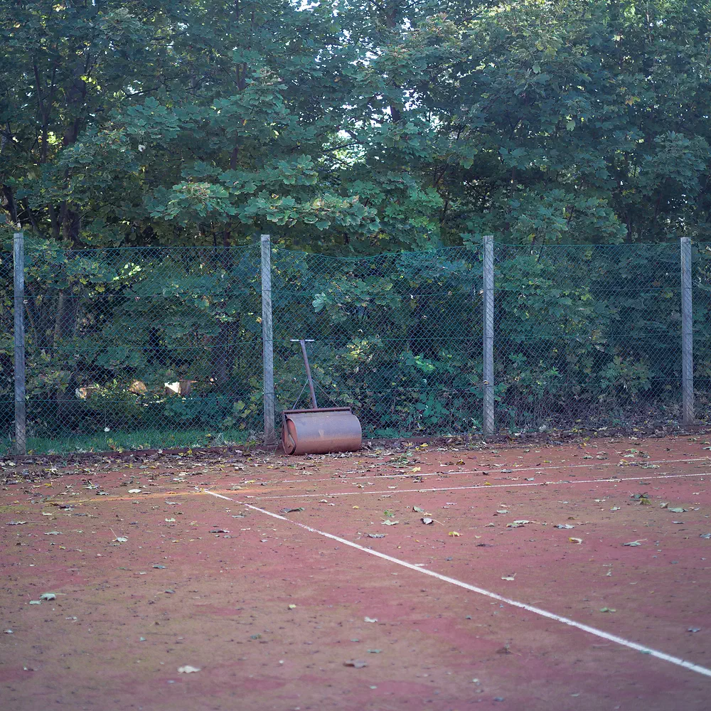 Overgrown clay tennis court with a roller in front of a fence and trees at Sjællands Odde