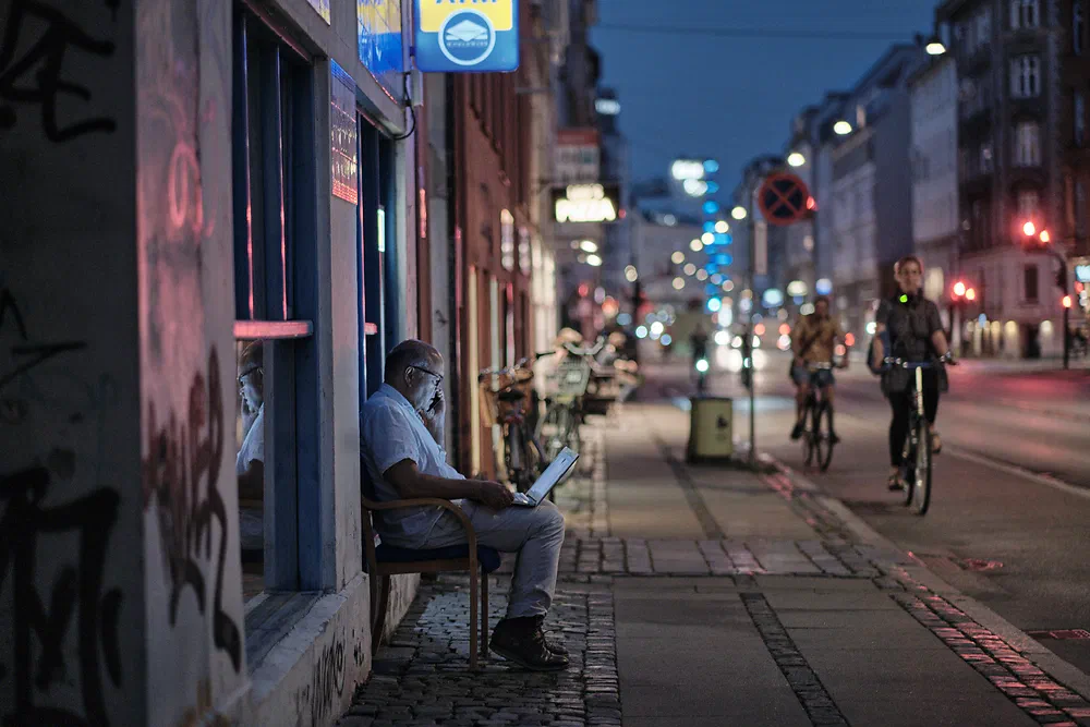 Man sitting on a bench with his laptop on a Vesterbro street at dusk while cyclists pass