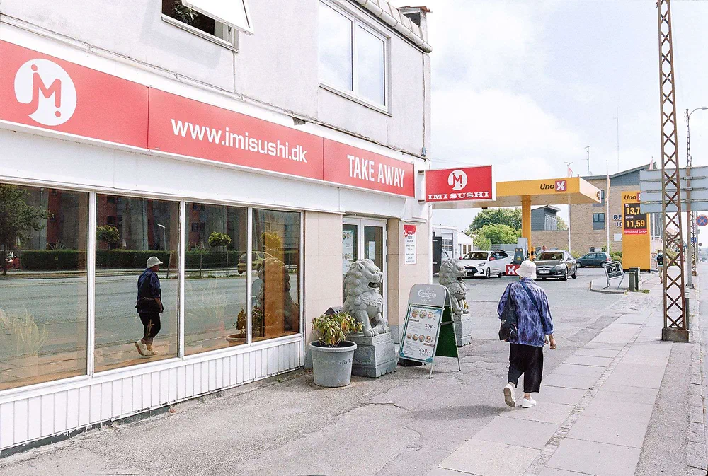 Woman in a sun hat walking past the Imi Sushi take away restaurant with stone lions on Roskildevej in Rødovre